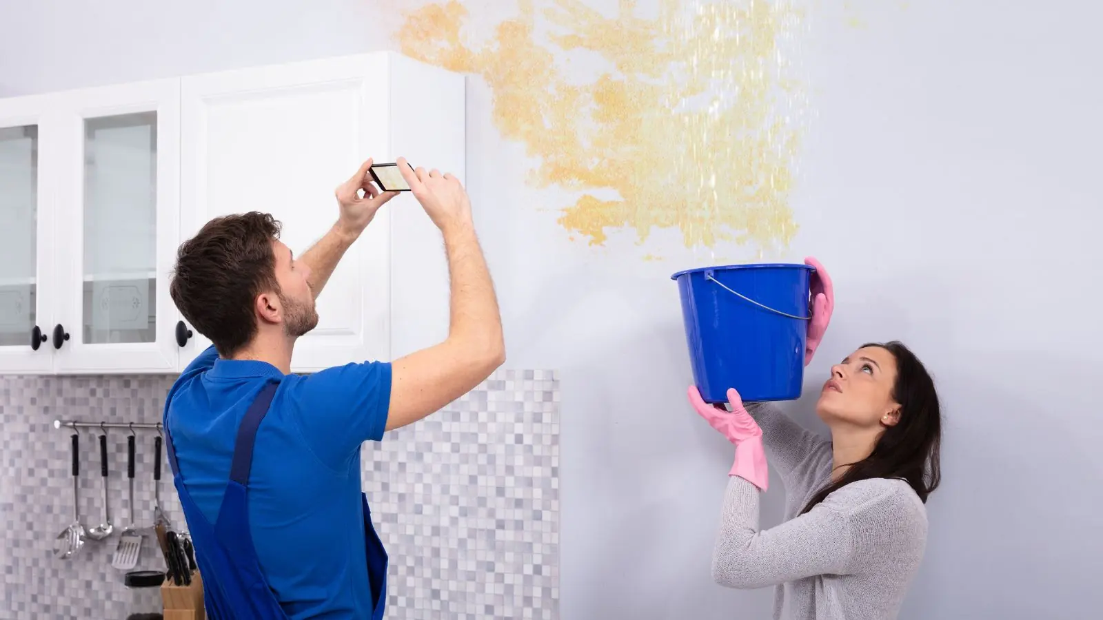 Homeowner holding a bucket under a ceiling leak while a technician photographs the damage
